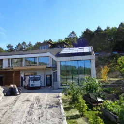 Nowoczesny dom z dużymi oknami w trakcie budowy, z widocznymi elementami konstrukcji i materiałami budowlanymi na podjeździe, otoczony zielenią i drzewami.