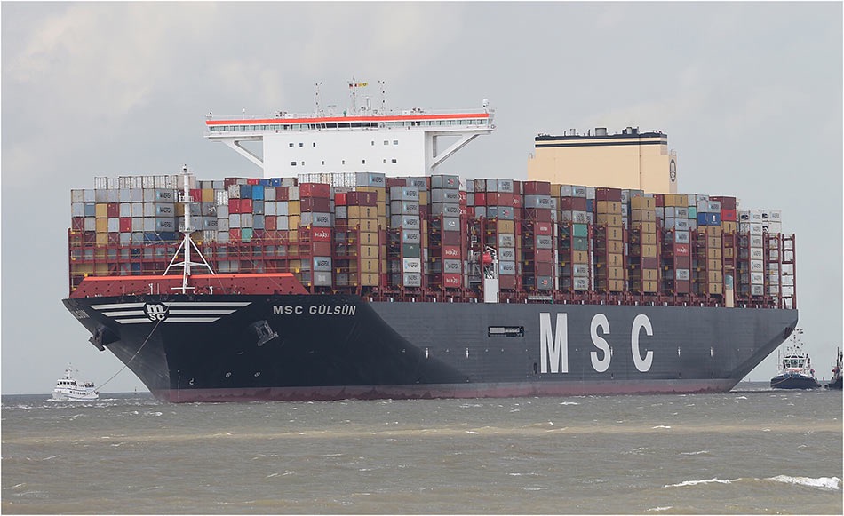 Ogromny kontenerowiec MSC Gülsün, załadowany wielobarwnymi kontenerami, płynie po wzburzonej wodzie, eskortowany przez dwa holowniki.