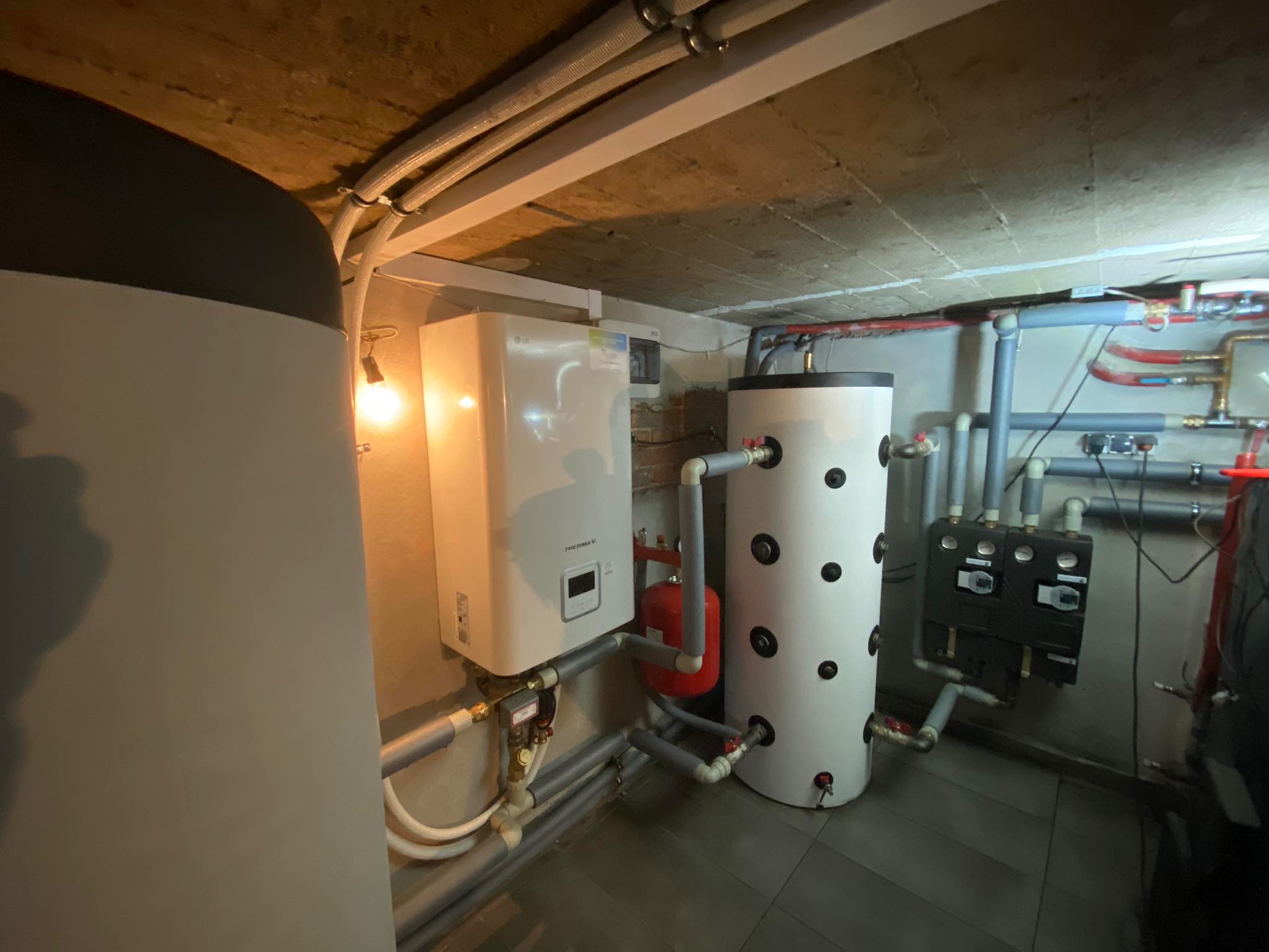 Zainstalowana pompa ciepła LG Therma V w piwnicy, widoczne rury, zbiornik buforowy i sterownik, surowy betonowy sufit z instalacjami.