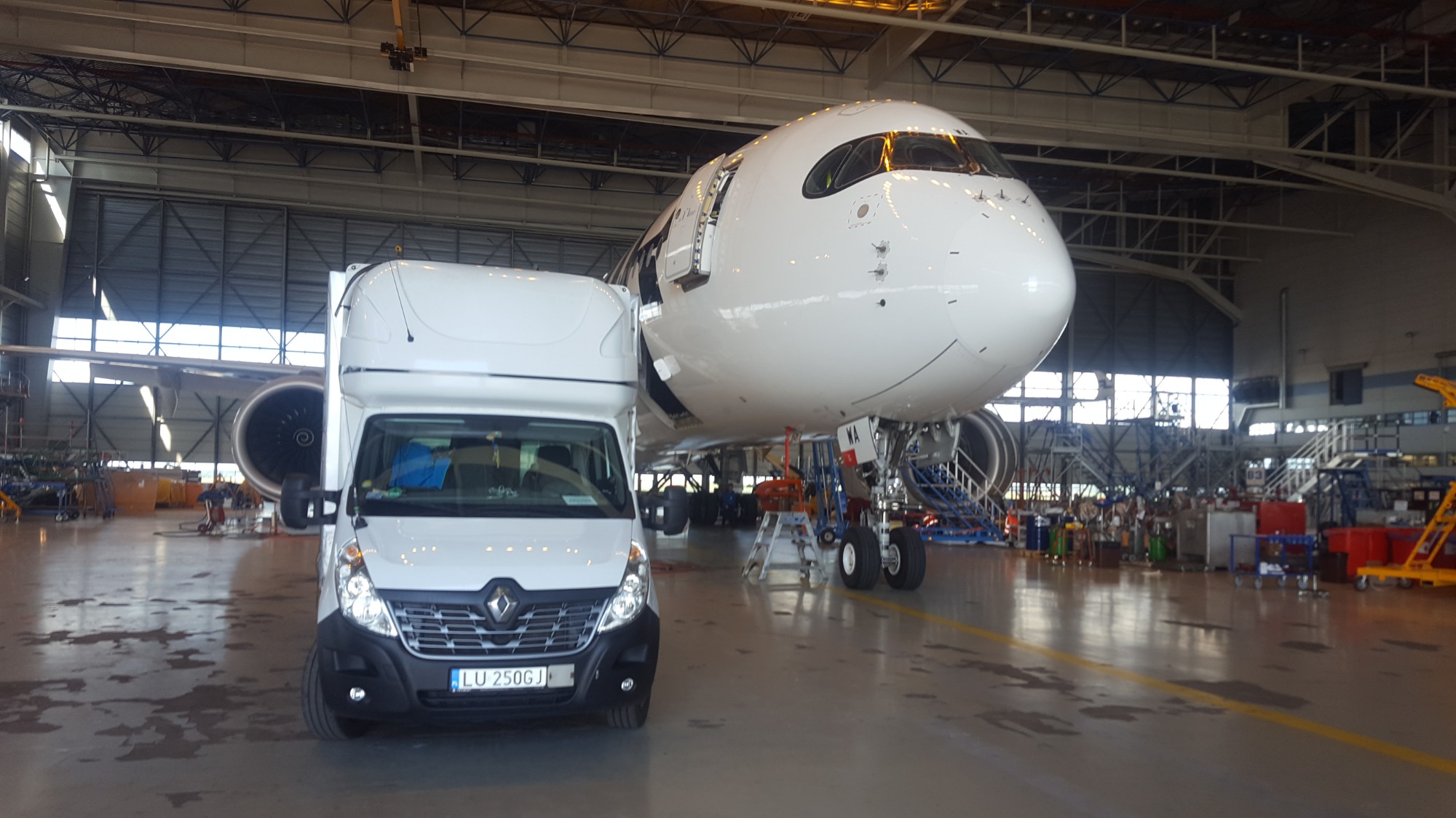 Biały bus Renault Master z wysoką kabiną zaparkowany obok ogromnego, białego samolotu pasażerskiego w hangarze lotniczym; widoczne turbiny samolotu, podwozie i schody; w tle konstrukcja hangaru.