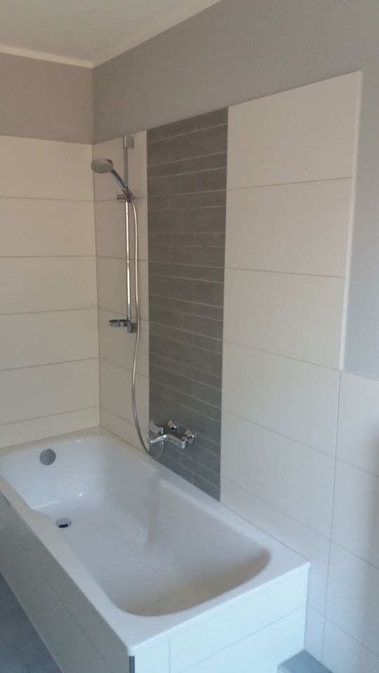 Biała wanna obudowana płytkami, ściany wyłożone białymi płytkami z szarym pasem z drobnej mozaiki, chromowana bateria i słuchawka prysznicowa.