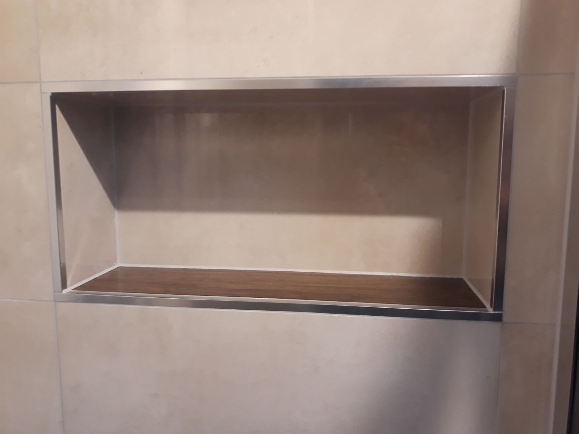 Wnęka w ścianie łazienki wyłożonej beżowymi płytkami, wykończona metalową ramką i półką z imitacji drewna.