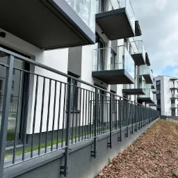 Nowoczesne balkony z szklanymi balustradami w nowym budownictwie, widoczne z poziomu gruntu, z niską, ciemną poręczą oddzielającą budynek od terenu zielonego.