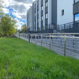 Zewnętrzna rampa z poziomymi, metalowymi poręczami prowadząca do nowoczesnego budynku z szaro-białą elewacją, otoczona zielonym trawnikiem i drzewami pod błękitnym, obłocznym niebem.