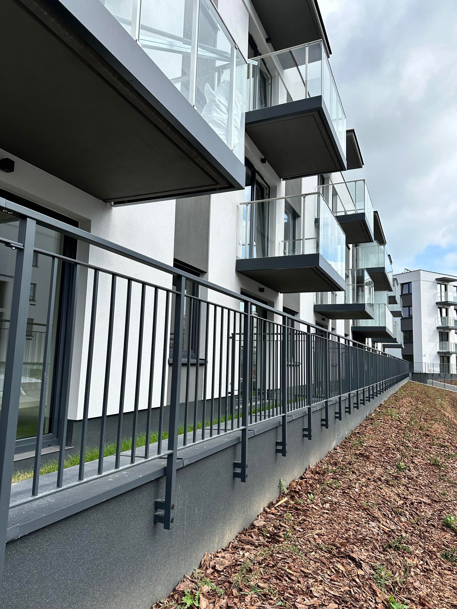 Nowoczesne balkony z szklanymi balustradami w nowym budownictwie, widoczne z poziomu gruntu, z niską, ciemną poręczą oddzielającą budynek od terenu zielonego.