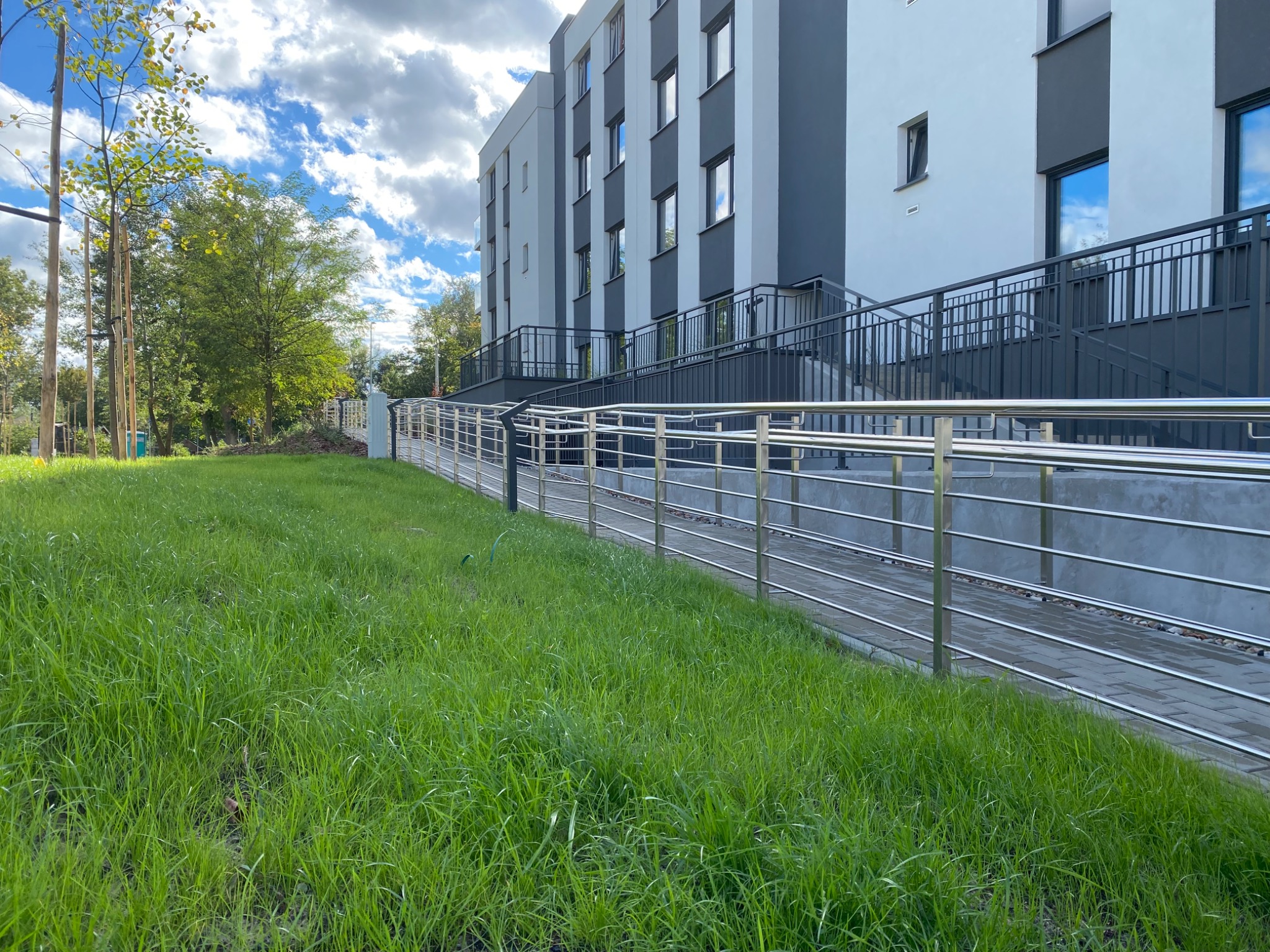Zewnętrzna rampa z poziomymi, metalowymi poręczami prowadząca do nowoczesnego budynku z szaro-białą elewacją, otoczona zielonym trawnikiem i drzewami pod błękitnym, obłocznym niebem.