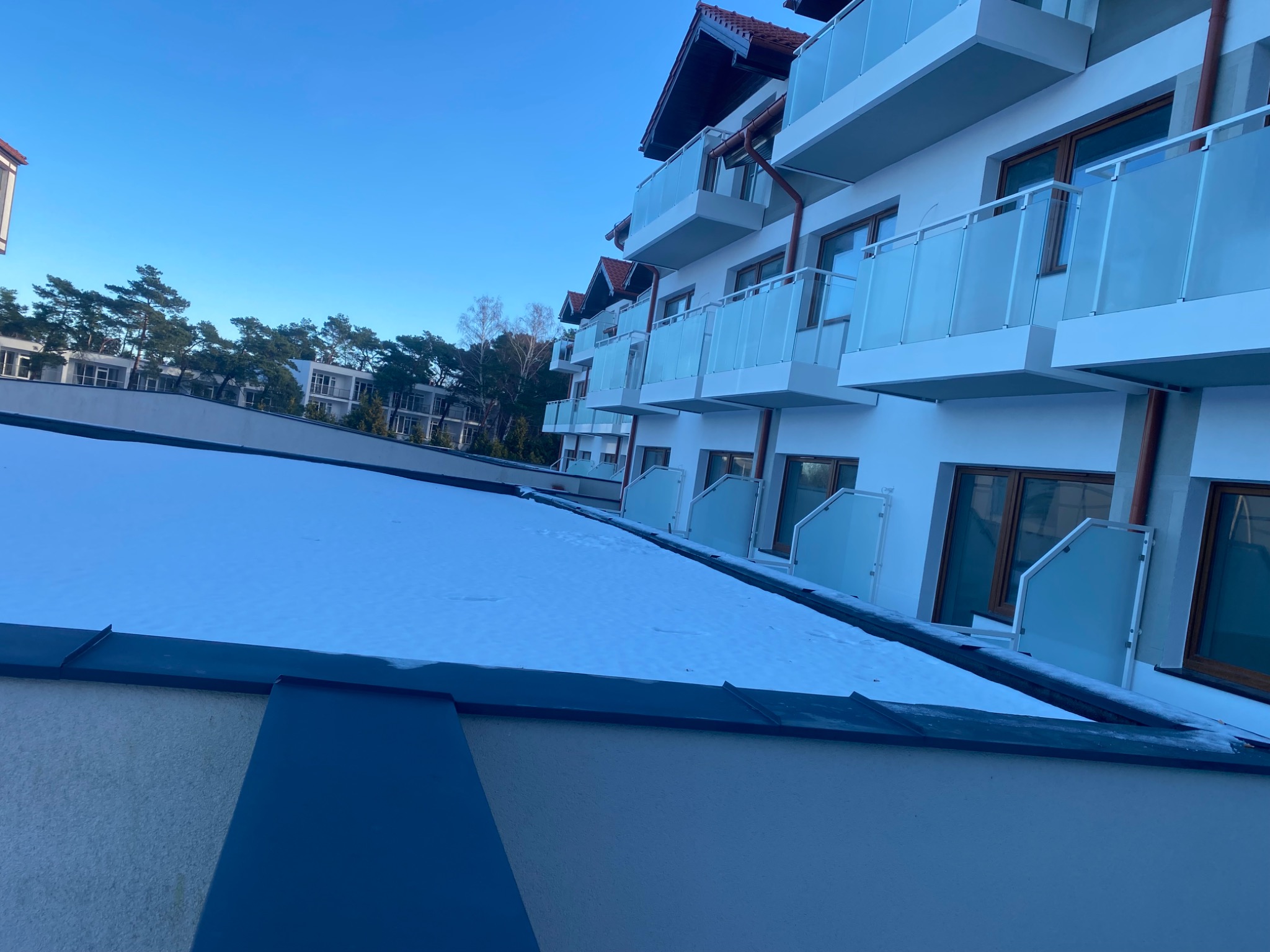 Nowoczesny budynek apartamentowy z balkonami zabezpieczonymi szkłem, widok z góry na zasypany śniegiem dach.