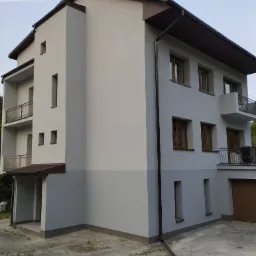 Dwukondygnacyjny dom z jasnoszarą elewacją, brązowymi oknami, balkonami z metalowymi balustradami i brązowym dachem, sfotografowany w pochmurny dzień.