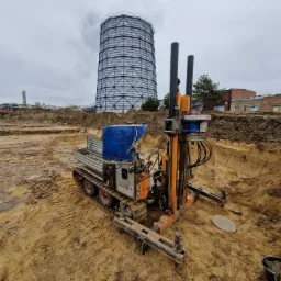 Wiertnica na gąsienicach podczas prac ziemnych we Wrocławiu, w tle widoczna chłodnia kominowa i wykop.