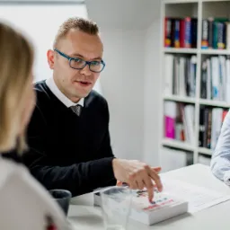 Mężczyzna w okularach tłumaczy tekst z książki 'Active English' dwóm kobietom przy stole, w tle regał z książkami.