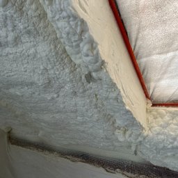 PAKRIS.pl Patryk Szydłowski - Świeżo nałożona, jasna pianka izolacyjna wypełnia przestrzeń poddasza, przylegając do czerwonej rurki i metalowej siatki, tworząc nierówną, porowatą strukturę.