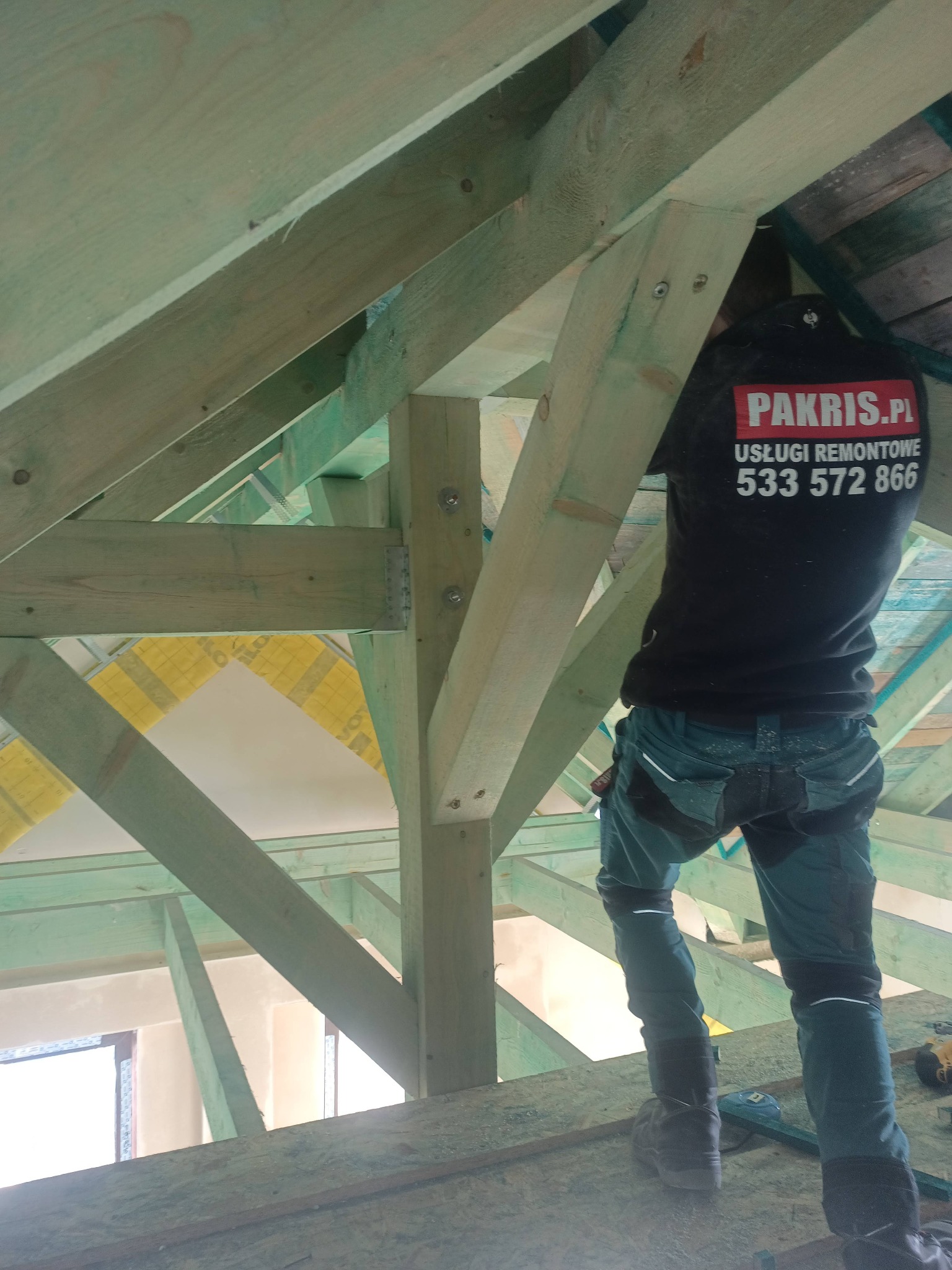 Prace ociepleniowe poddasza: widoczne drewniane elementy konstrukcyjne dachu, żółta folia izolacyjna i pracownik w uniformie z logo firmy Pakris.pl.