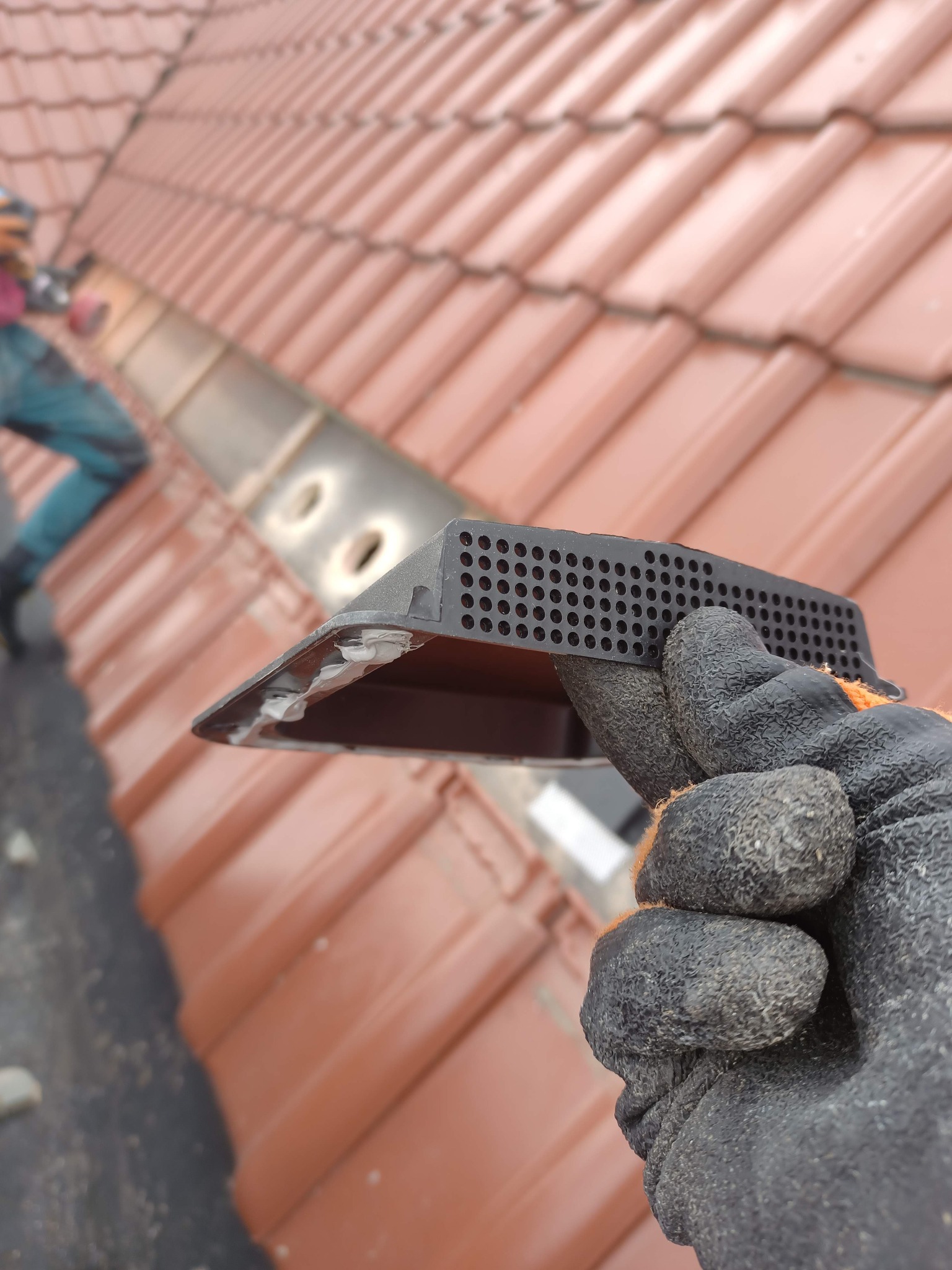 Montaż elementu wentylacyjnego dachu w Dobrym Mieście. Ręka w rękawicy trzyma czarny element z otworami na tle czerwonej dachówki. W tle pracownik podczas montażu.