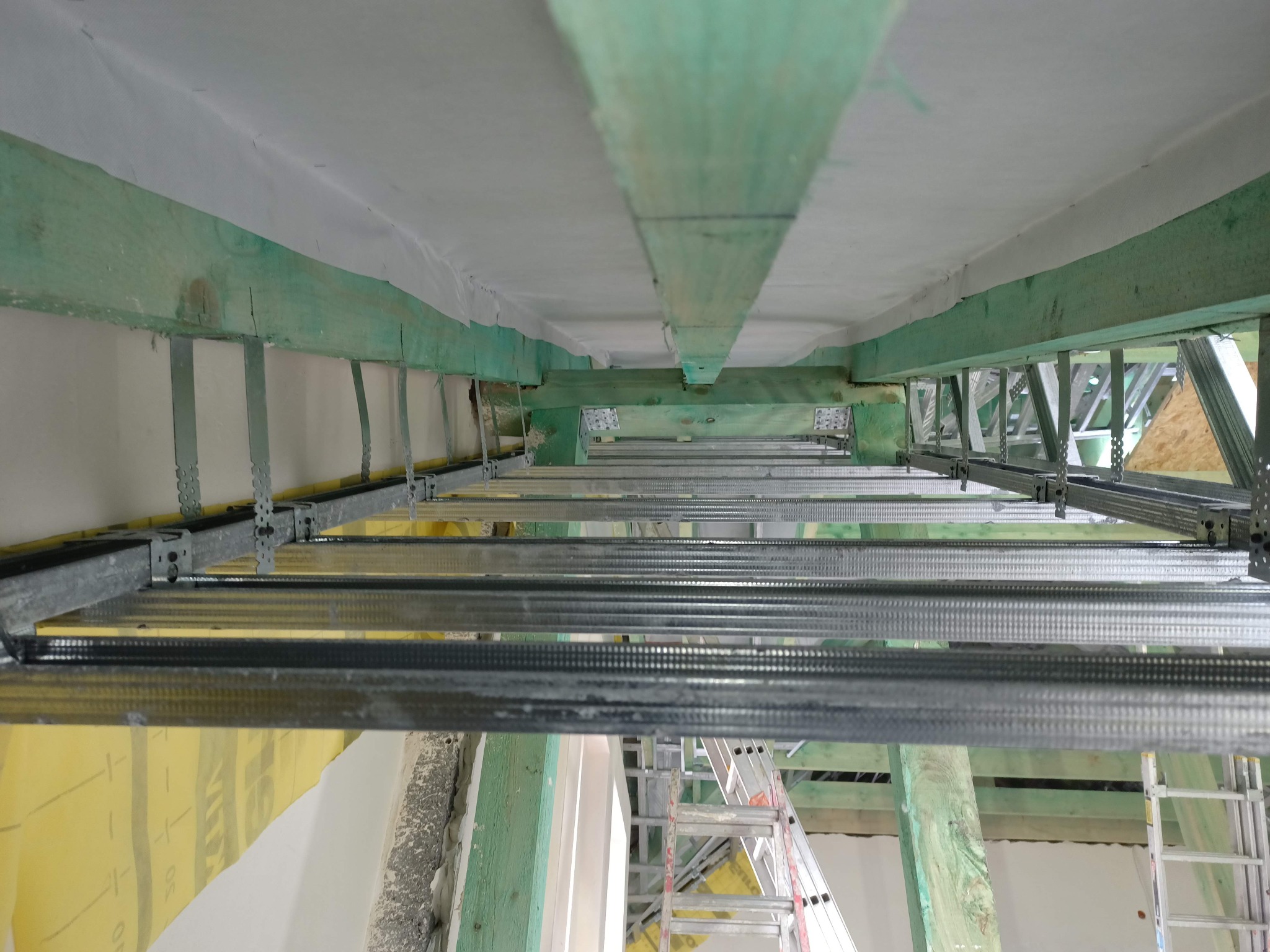 Sufit z drewnianą konstrukcją i metalowymi profilami, przygotowany do izolacji. Widoczna drabina i żółta folia izolacyjna.