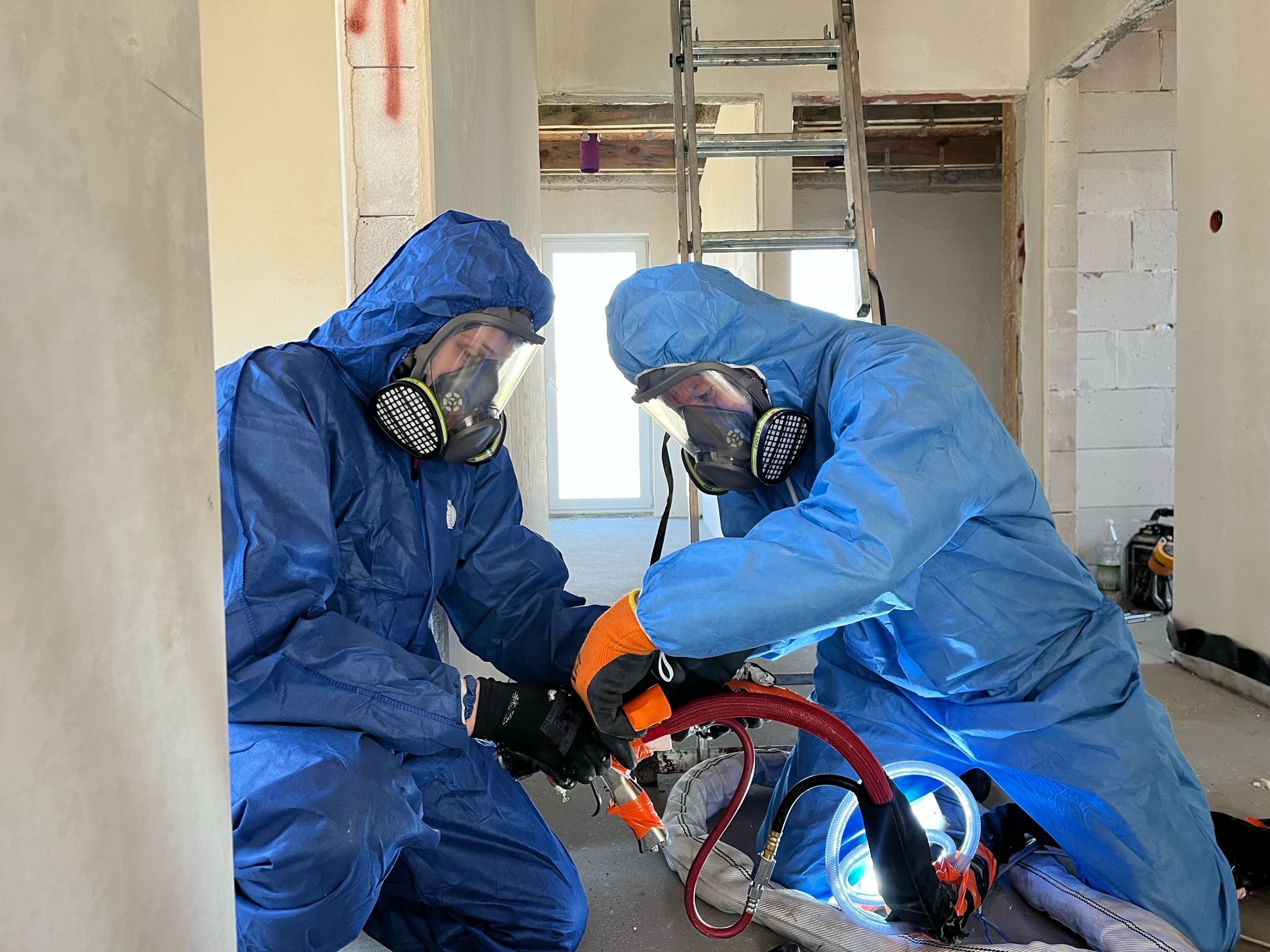 Pracownicy w kombinezonach ochronnych i maskach aplikują pianę, widoczna instalacja i drabina w surowym wnętrzu budynku w trakcie ocieplania.