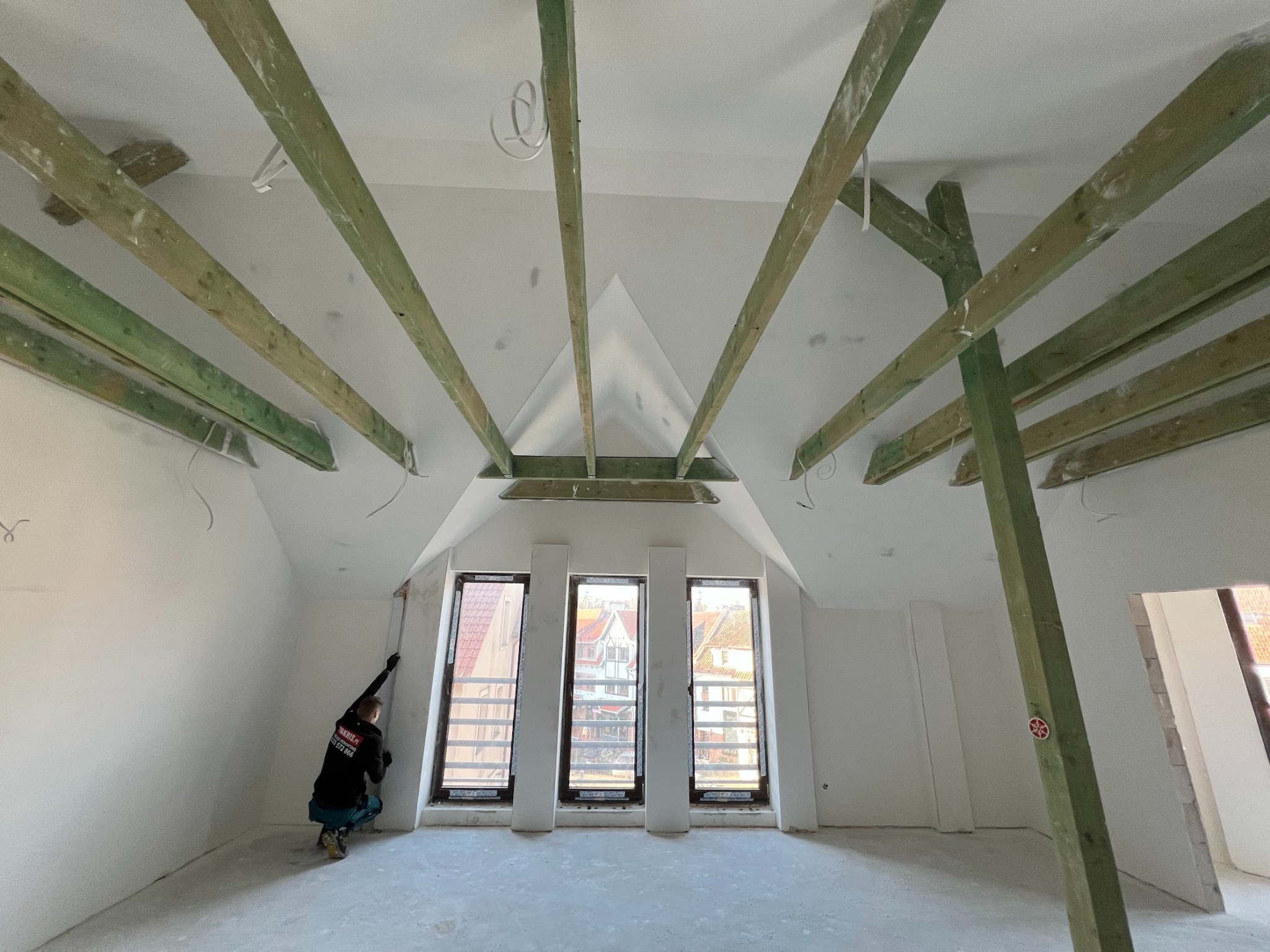 Prace wykończeniowe poddasza: montaż izolacji przy oknach dachowych. Widoczne drewniane belki konstrukcyjne i pracownik w trakcie pracy. W tle zabudowa okien.