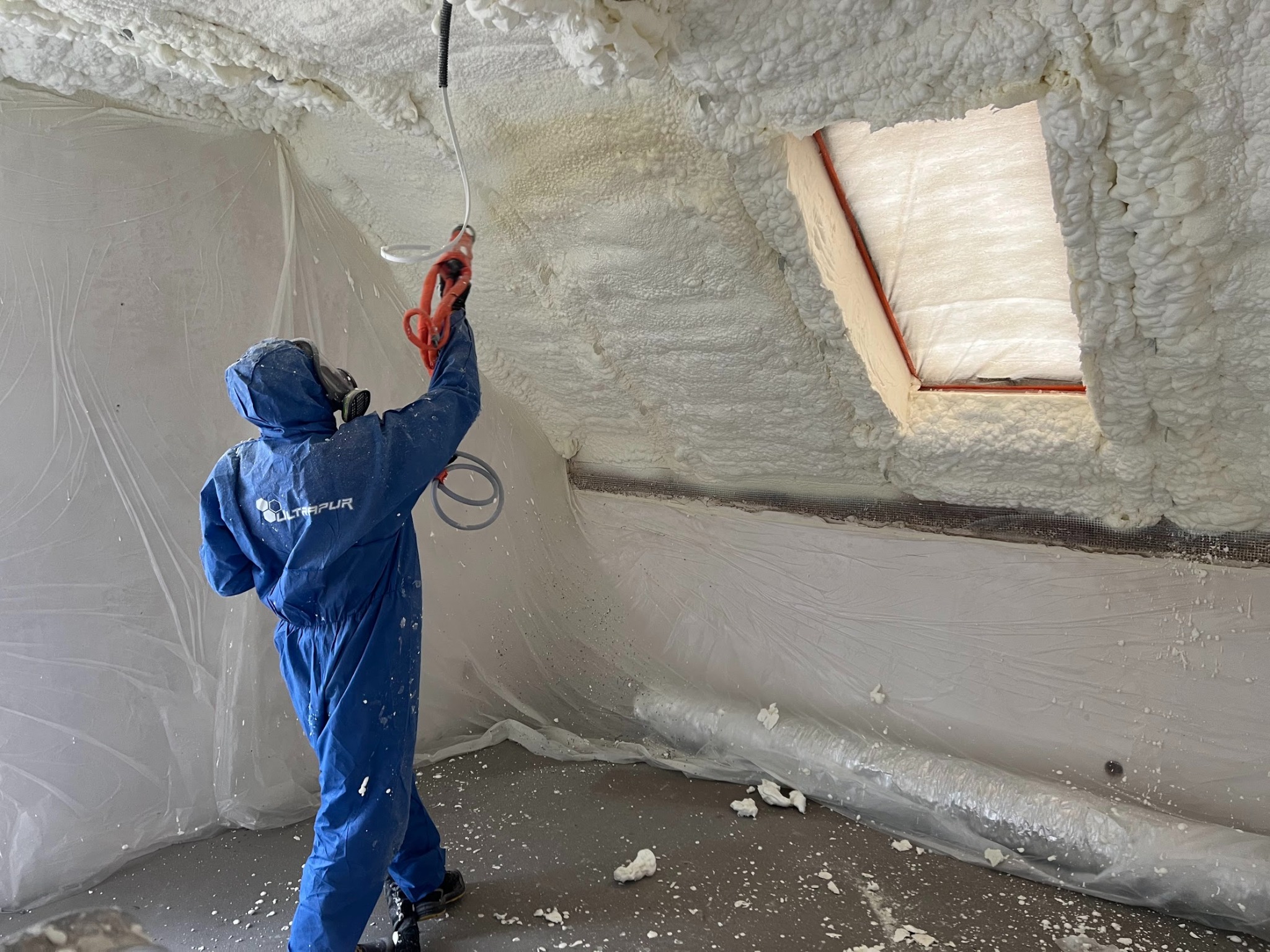 Pracownik w kombinezonie ochronnym aplikuje pianę izolacyjną na poddaszu, widoczne okno dachowe i folia ochronna na ścianach. Detal: logo firmy na kombinezonie.