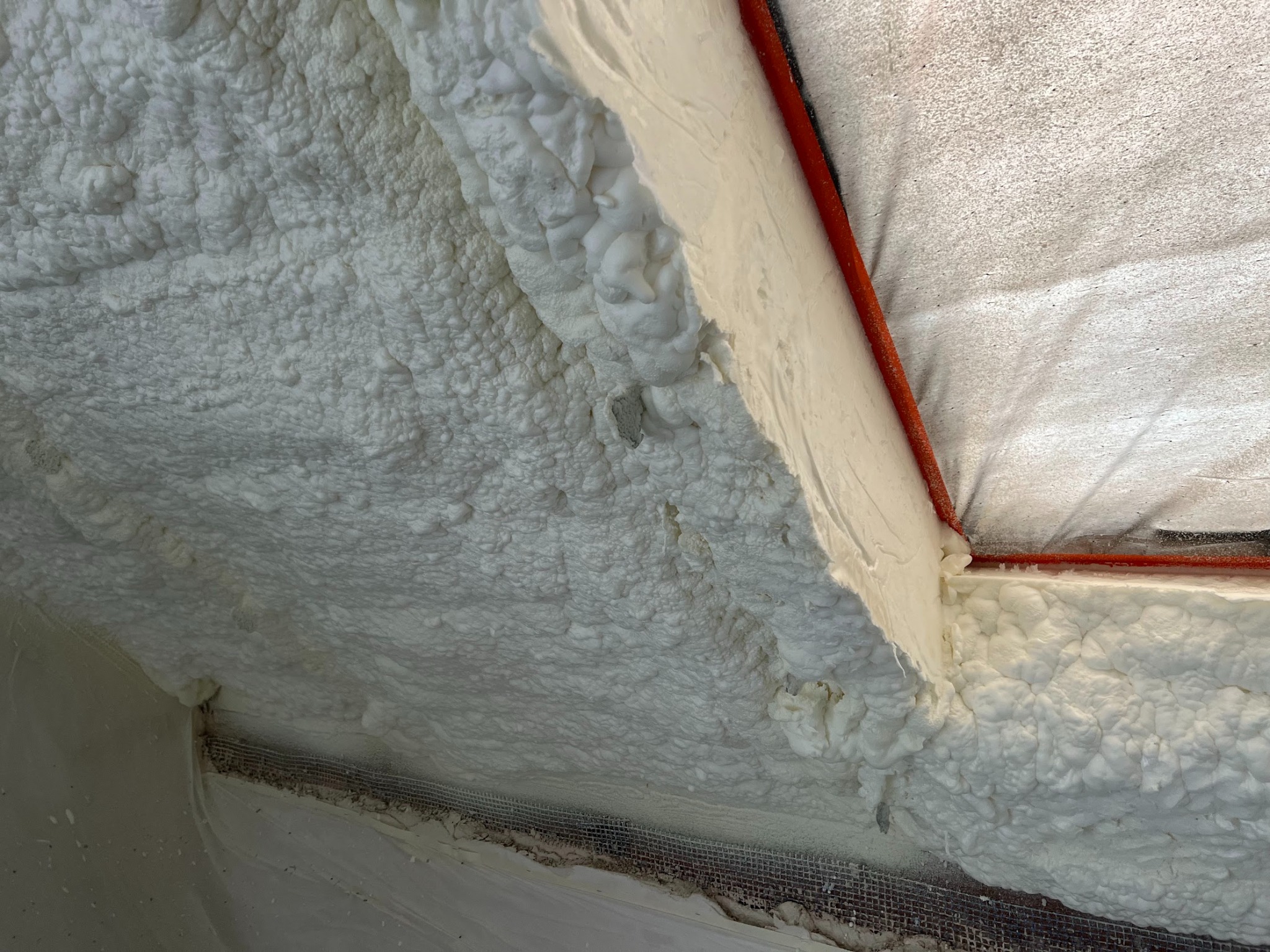 Świeżo nałożona, jasna pianka izolacyjna wypełnia przestrzeń poddasza, przylegając do czerwonej rurki i metalowej siatki, tworząc nierówną, porowatą strukturę.