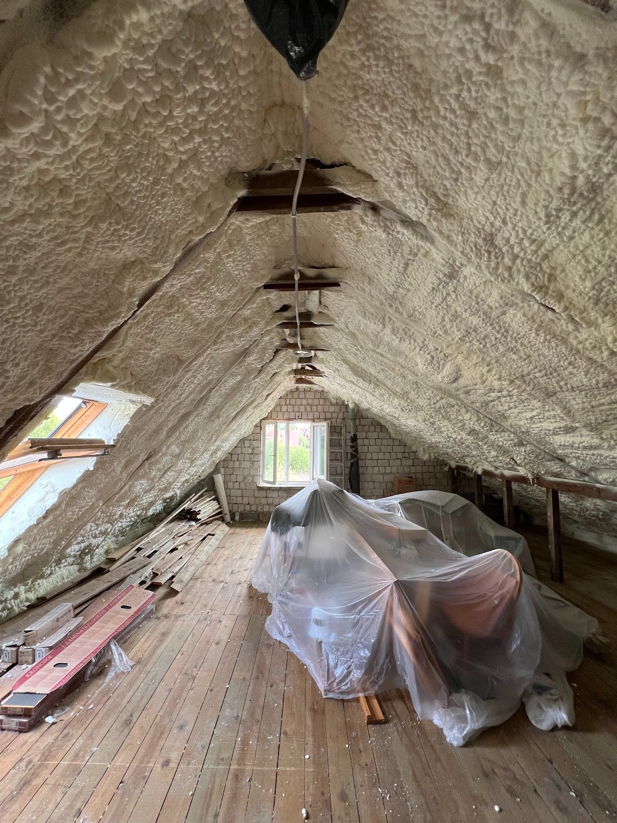 Poddasze po natrysku izolacji. Pianka pokrywa skośne ściany i sufit. Okno w szczycie, podłoga z desek, meble zabezpieczone folią. Widoczne drewniane elementy konstrukcyjne.