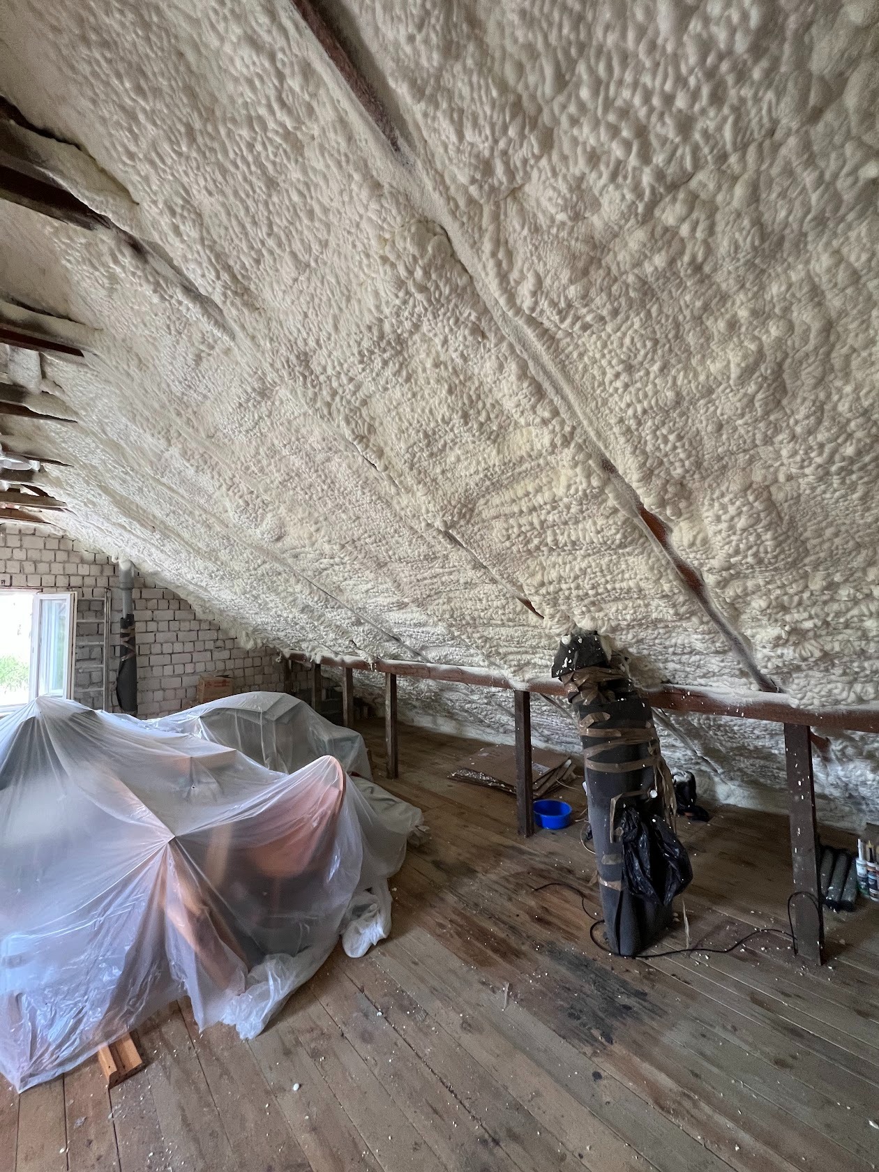 Ocieplone poddasze pianką, widoczne krokwie. Meble zabezpieczone folią. Izolacja natryskowa na skośnym dachu. Prace ociepleniowe na poddaszu z drewnianą podłogą.