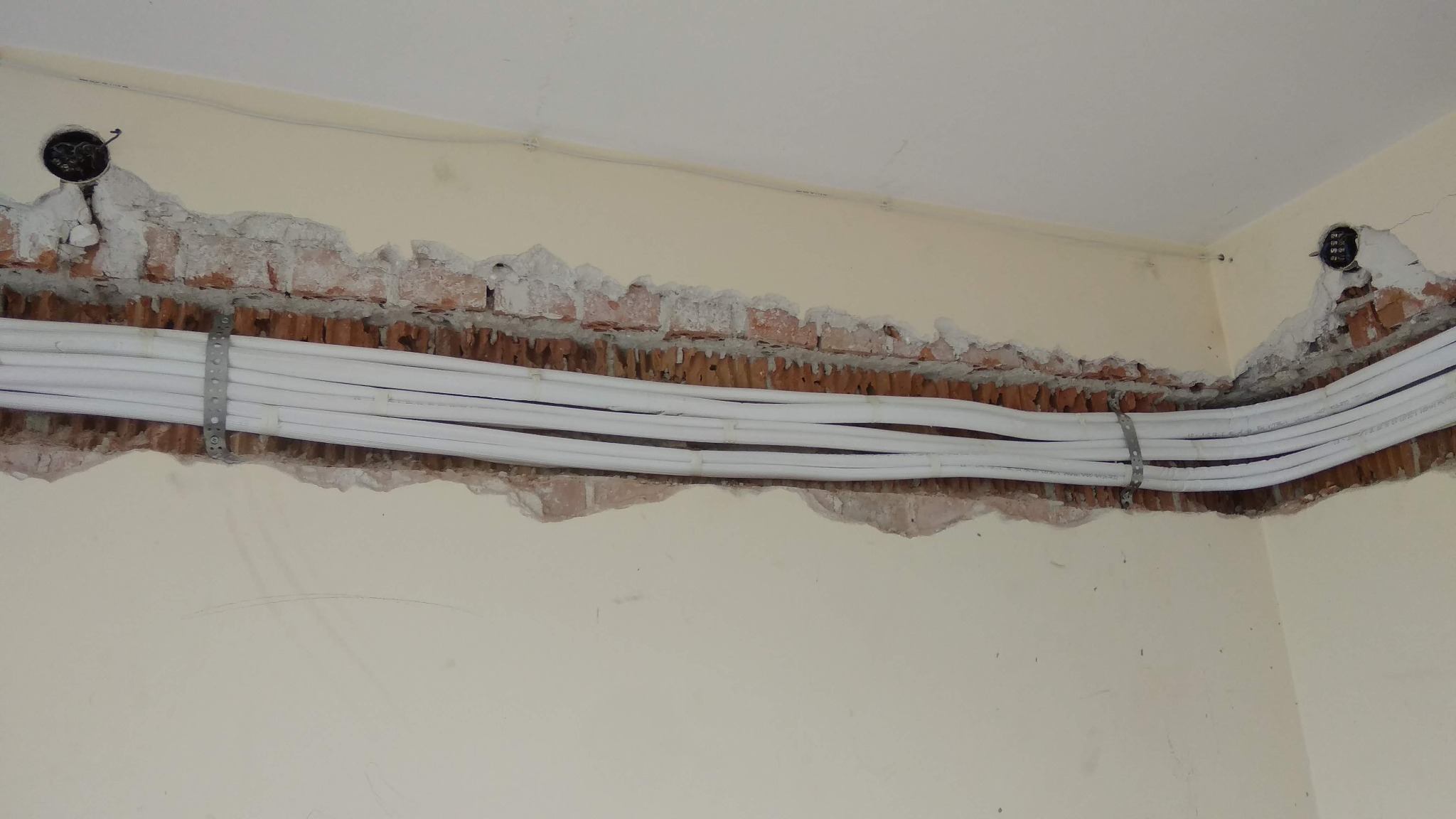 Białe rury instalacyjne, przymocowane metalowymi obejmami do ściany z widoczną cegłą w bruździe, przygotowane pod tynkowanie w domu jednorodzinnym.