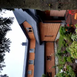 Dom z drewnianą elewacją i ciemnym dachem z blachy trapezowej, widok z poziomu ogrodu z roślinami i kwiatami