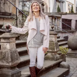 Uśmiechnięta blondynka w jasnej stylizacji (pudrowy róż, biel, szarość) pozuje na kamiennych schodach z ozdobną balustradą, spoglądając w górę