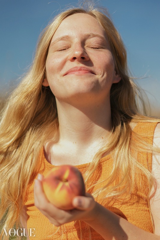 Portret kobiety z blond włosami, z zamkniętymi oczami i uśmiechem, trzymającej brzoskwinię na tle błękitnego nieba.
