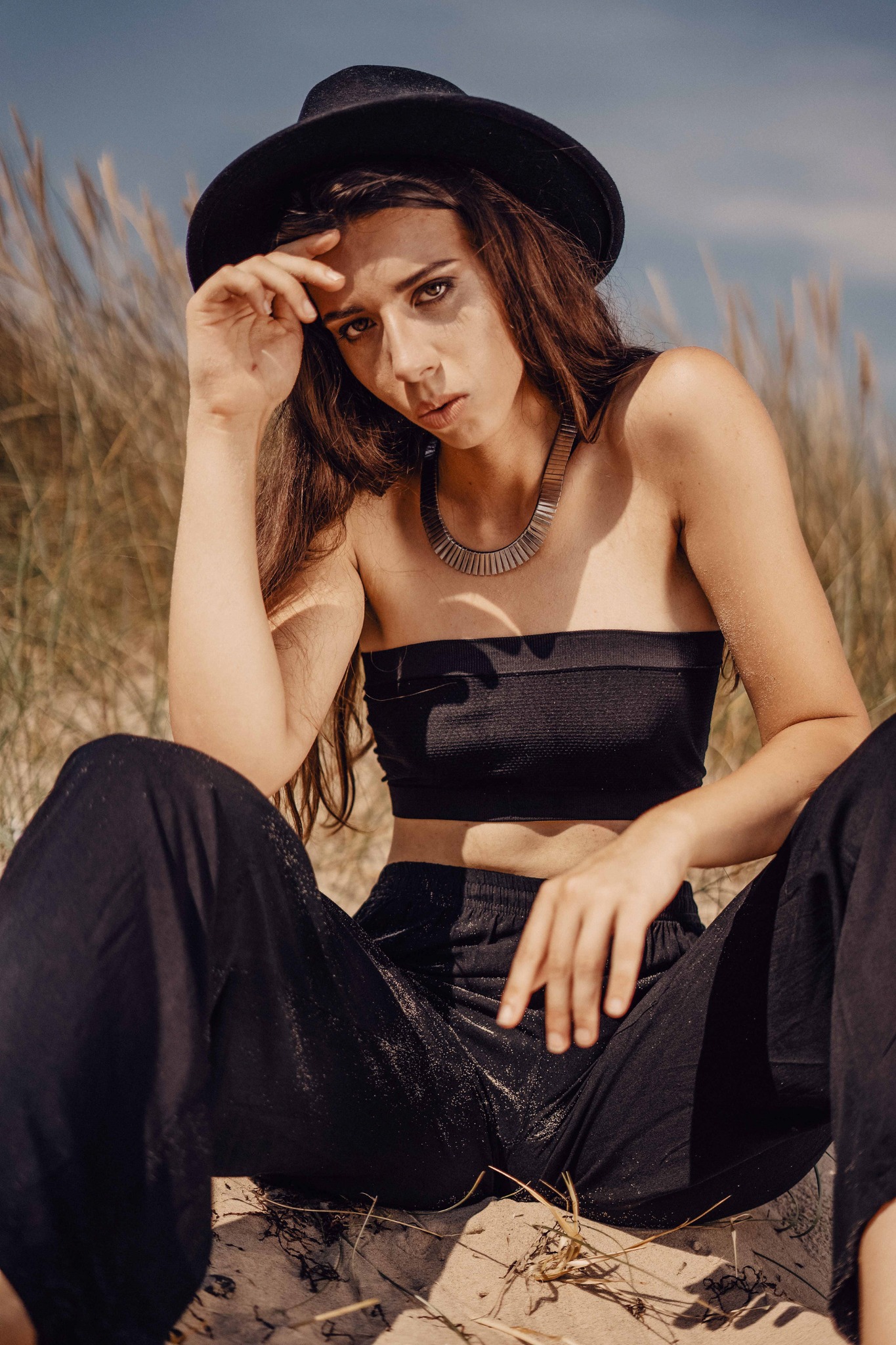 Portret kobiety w czarnym kapeluszu i topie, siedzącej na piasku z trawami w tle, z metalowym naszyjnikiem i lekkim makijażem.