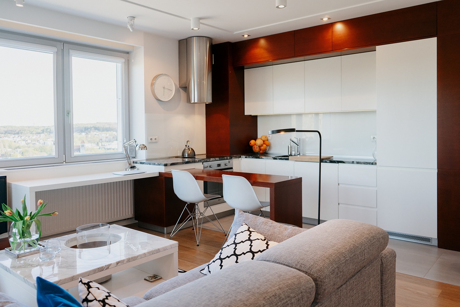 Nowoczesne wnętrze mieszkania z widokiem na miasto, obejmujące białą kuchnię z drewnianymi akcentami, marmurowy blat, stół z krzesłami, oraz część wypoczynkową z szarą sofą i marmurowym stolikiem...