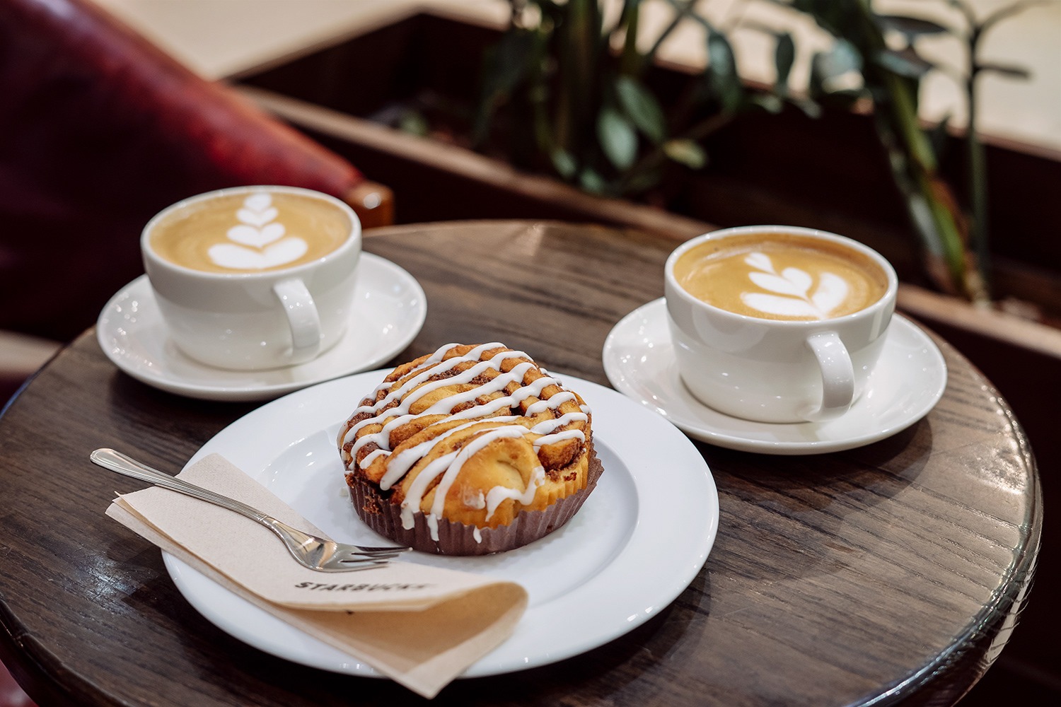 Dwa cappuccino z wzorem na piance i cynamonka z lukrem na białym talerzyku z serwetką i widelcem, na okrągłym drewnianym stoliku.