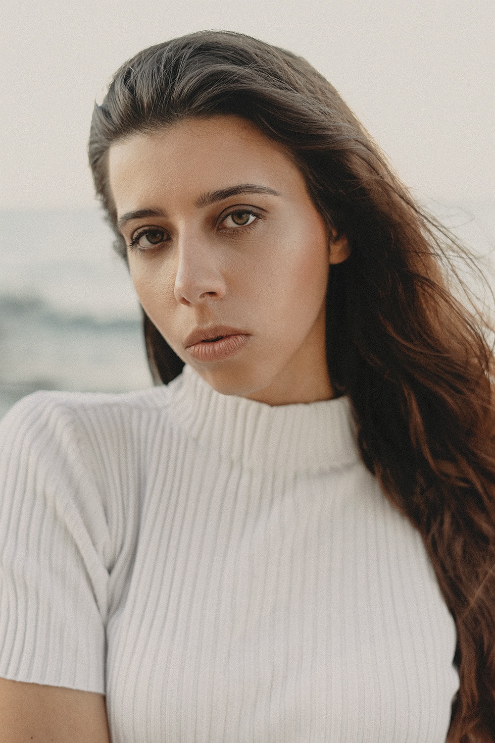 Portret kobiety o długich brązowych włosach i brązowych oczach, ubrana w biały sweter z golfem, spogląda wprost w obiektyw na tle rozmytego krajobrazu.