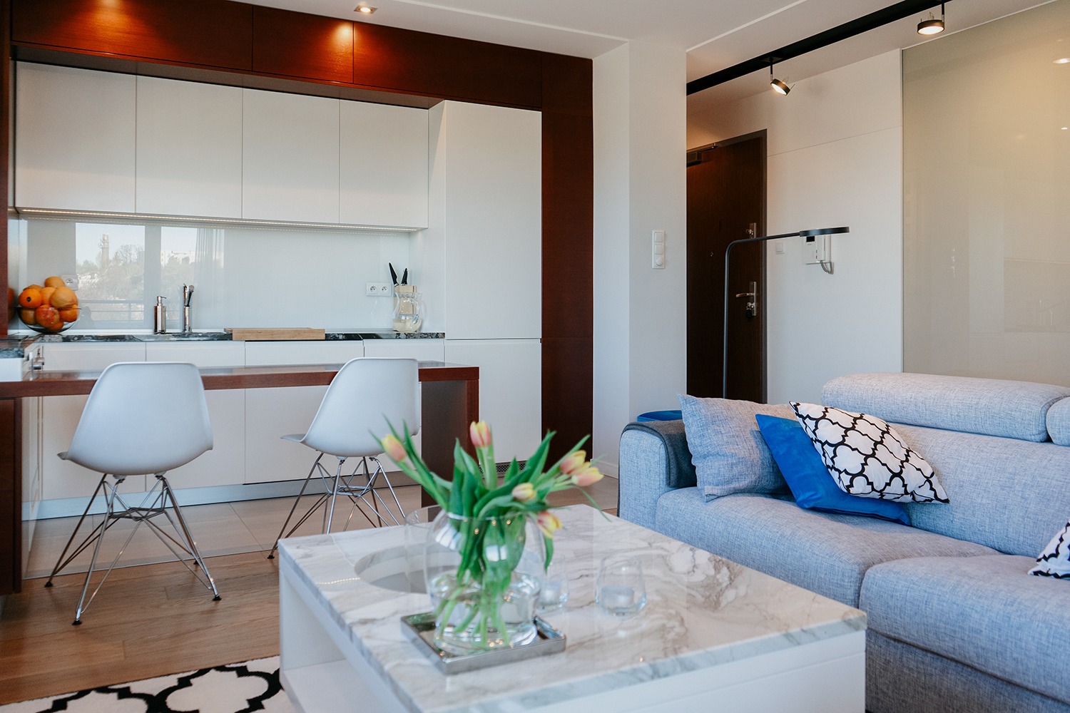 Jasne, nowoczesne wnętrze salonu z aneksem kuchennym: marmurowy stolik kawowy z tulipanami, szara sofa z dekoracyjnymi poduszkami, białe krzesła barowe przy blacie, minimalistyczna zabudowa...