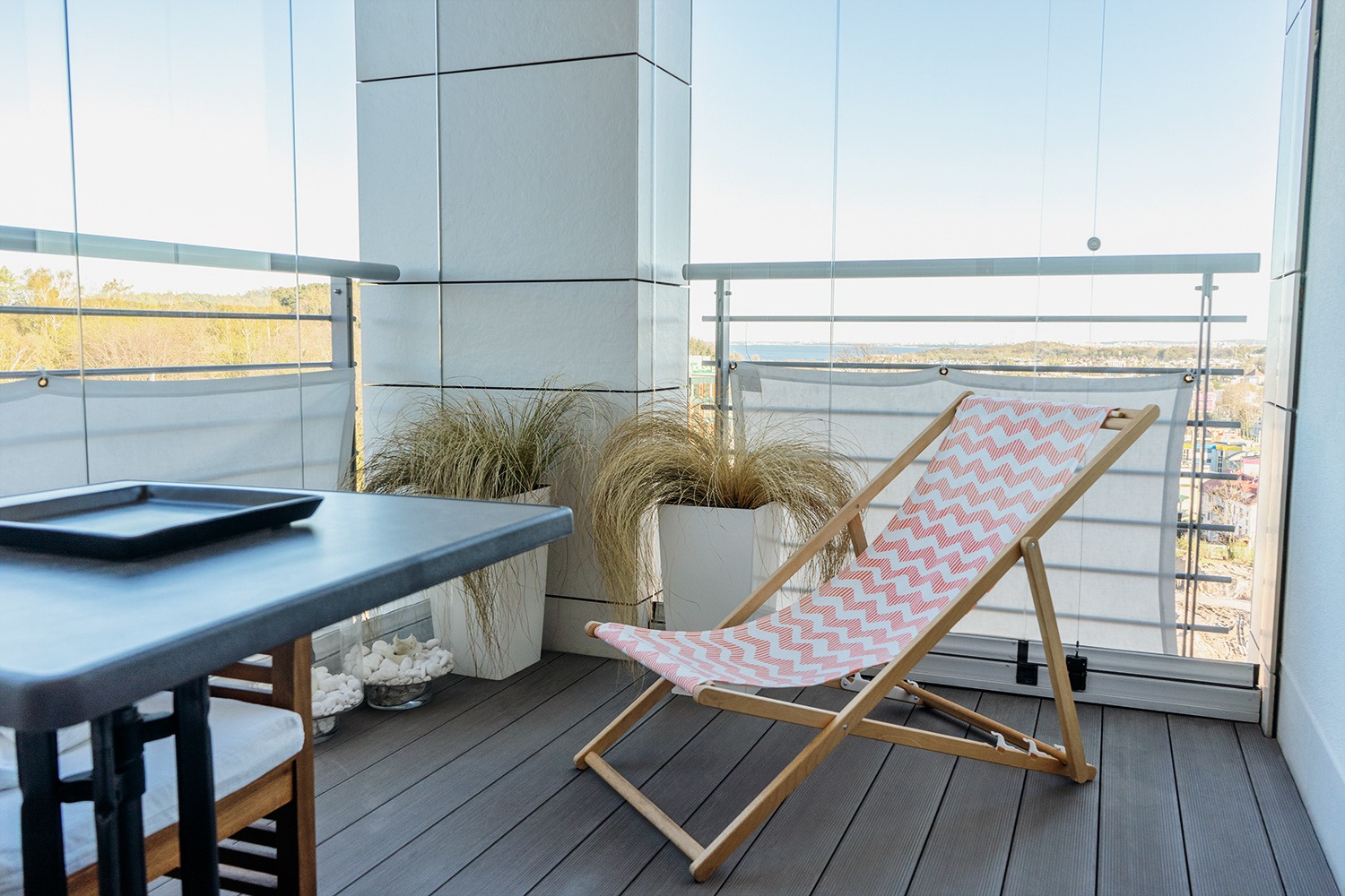 Nowoczesny balkon z drewnianą podłogą, szklaną balustradą, leżakiem w zygzaki, stolikiem z tacą i dekoracyjnymi trawami w białych donicach.