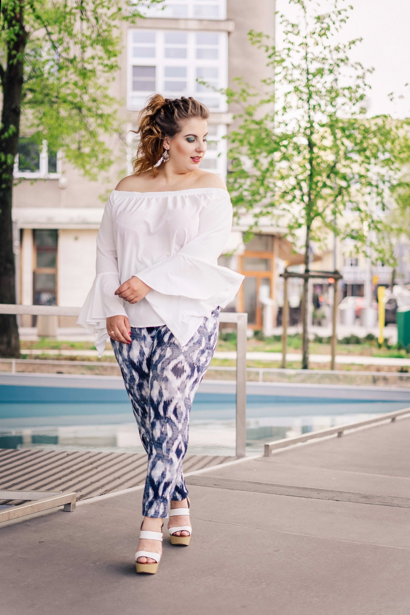 Kobieta w białej bluzce z odkrytymi ramionami i wzorzystych spodniach spaceruje w plenerze, z budynkiem w tle.