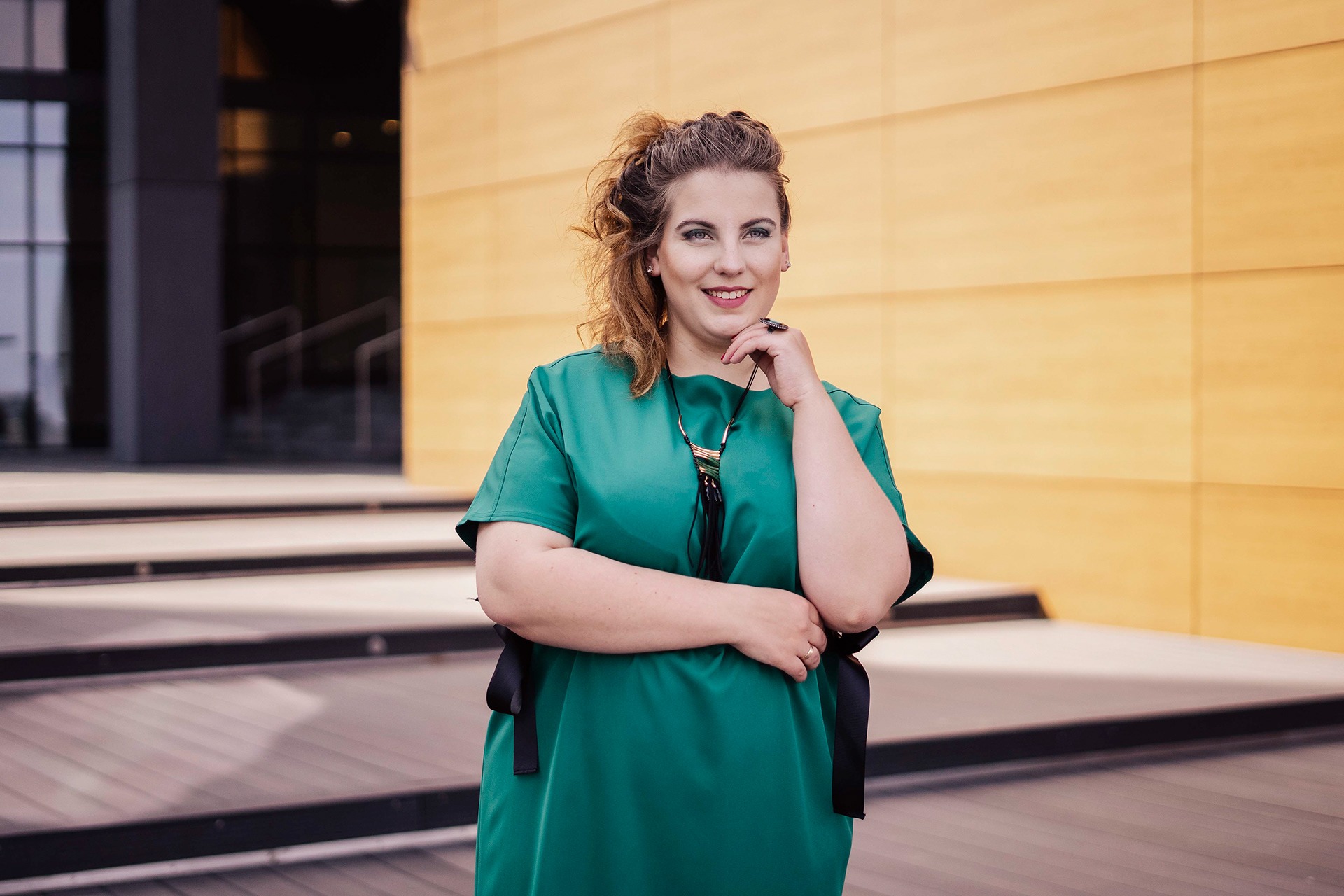Uśmiechnięta kobieta w szmaragdowej sukience z czarnymi akcentami i naszyjnikiem, pozująca na tle nowoczesnego budynku z poziomymi liniami.