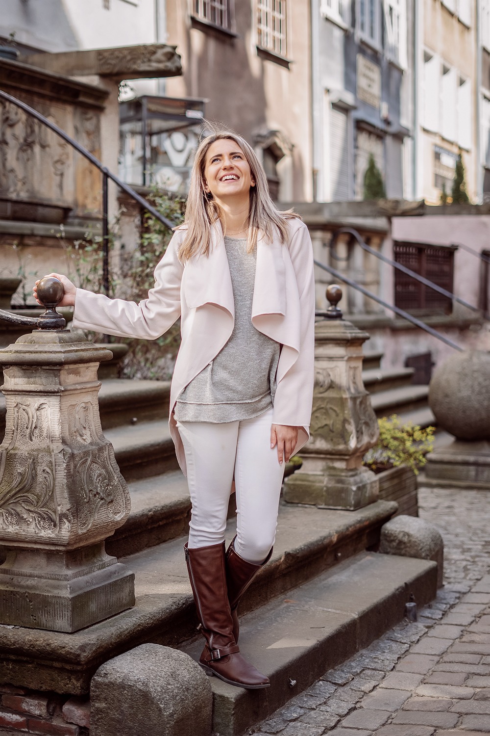 Uśmiechnięta blondynka w jasnej stylizacji (pudrowy róż, biel, szarość) pozuje na kamiennych schodach z ozdobną balustradą, spoglądając w górę