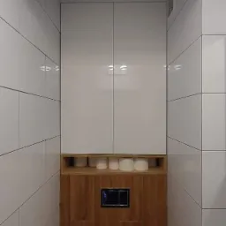 Nowoczesna łazienka po remoncie: białe, prostokątne płytki, zabudowa WC z drewnopodobną okładziną, szafka z połyskiem i zapas papieru toaletowego.