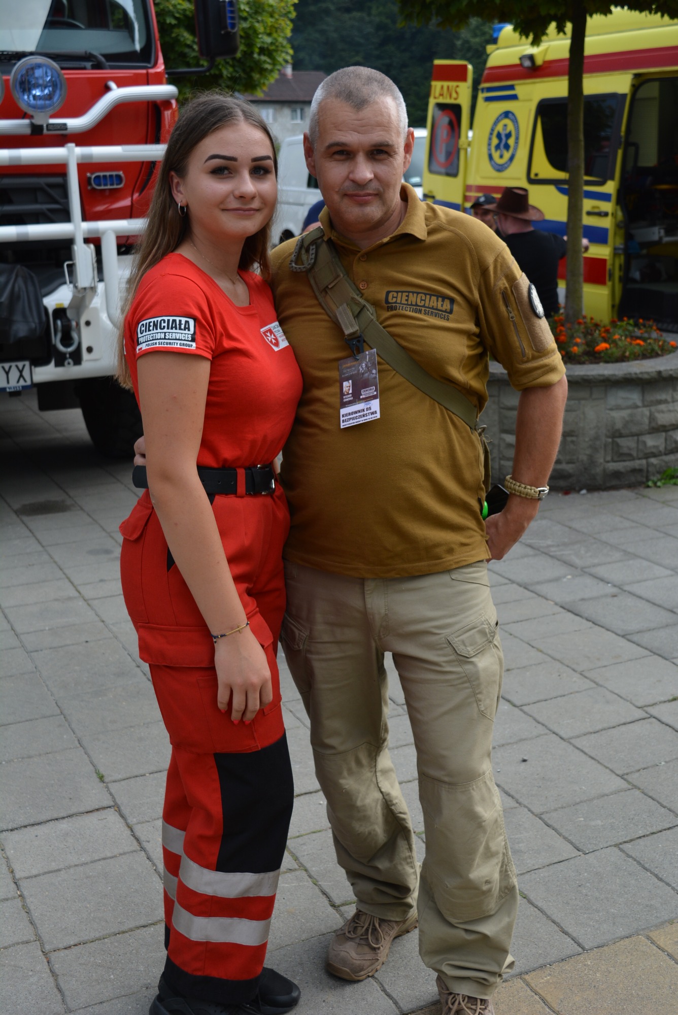 Mężczyzna i kobieta w uniformach z logo firmy, stojący na tle wozów strażackich i ambulansu.