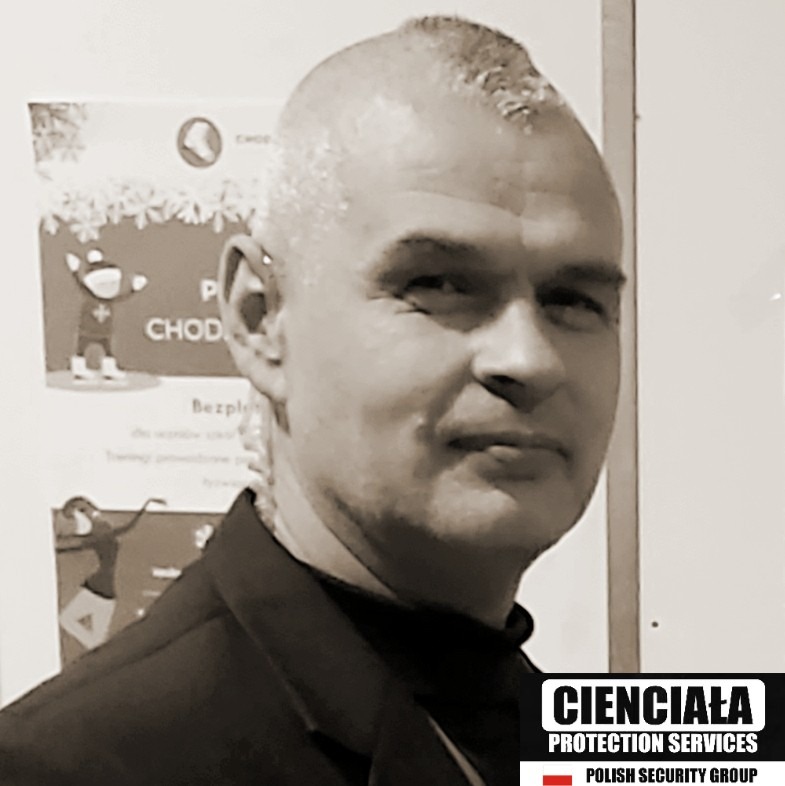 Portret mężczyzny w czarnej marynarce, z krótkimi włosami, z plakatem w tle i logo firmy ochroniarskiej w rogu.
