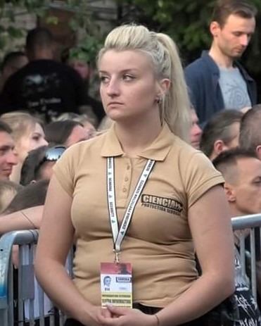 Blondwłosa kobieta w beżowej koszulce polo z identyfikatorem na smyczy, stojąca na tle tłumu ludzi i metalowej barierki.