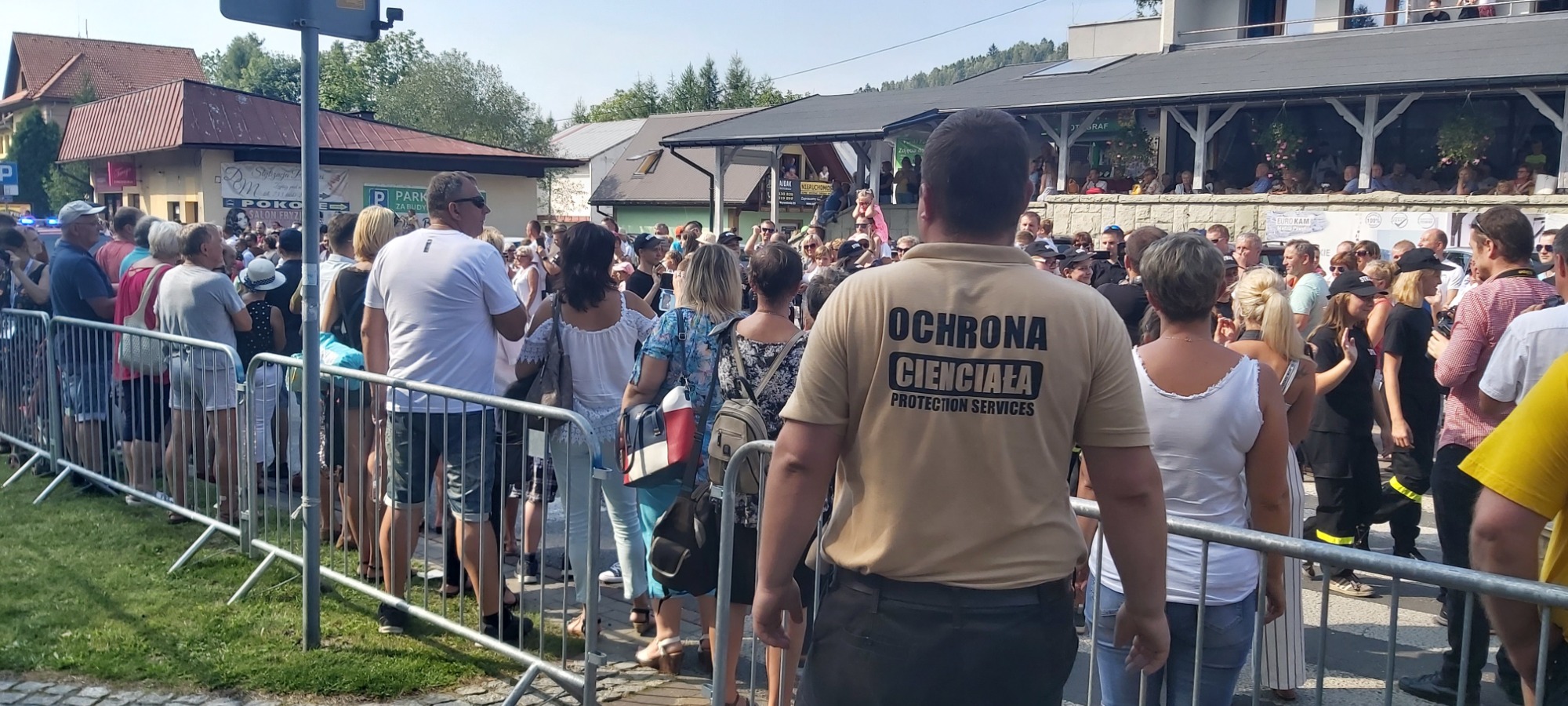 Ochroniarz w beżowej koszulce z napisem 'OCHRONA CIENCIAŁA PROTECTION SERVICES' stoi tyłem do kamery, obserwując tłum ludzi za metalowymi barierkami podczas wydarzenia plenerowego.