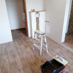 Wnętrze łazienki w trakcie remontu: drewnopodobne płytki podłogowe, drabina malarska, wiadro glazurnicze z gąbką, częściowo wykończona ściana z białych płytek i dekoracyjną mozaiką, widoczne...