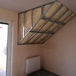 Metalowa konstrukcja pod zabudowę skośnego poddasza z widoczną izolacją z pianki, grzejnik i surowa ściana w trakcie prac wykończeniowych.