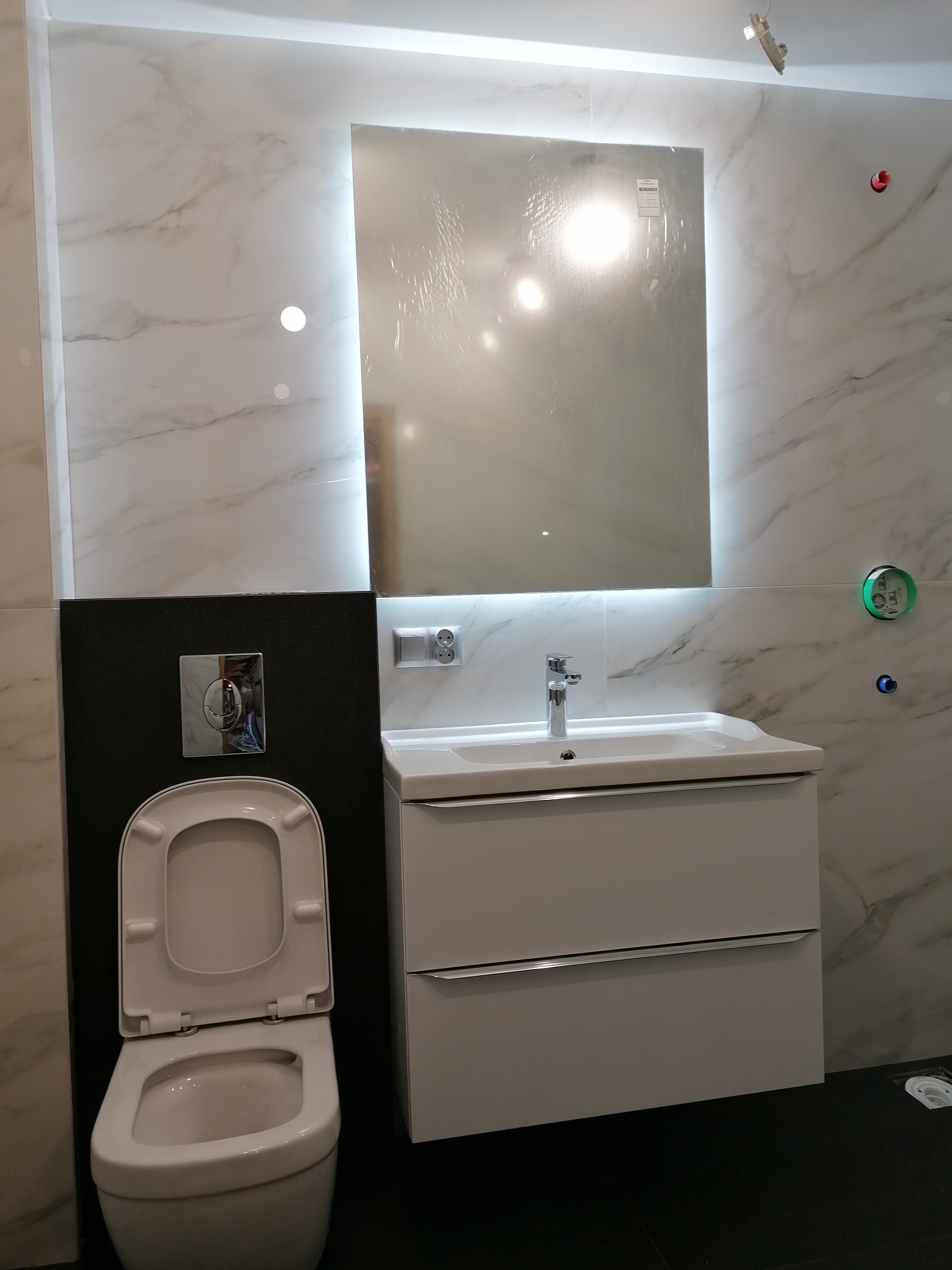 Nowoczesna łazienka z marmurowymi płytkami, podświetlanym lustrem, wiszącą toaletą i szafką pod umywalką z chromowanymi uchwytami.