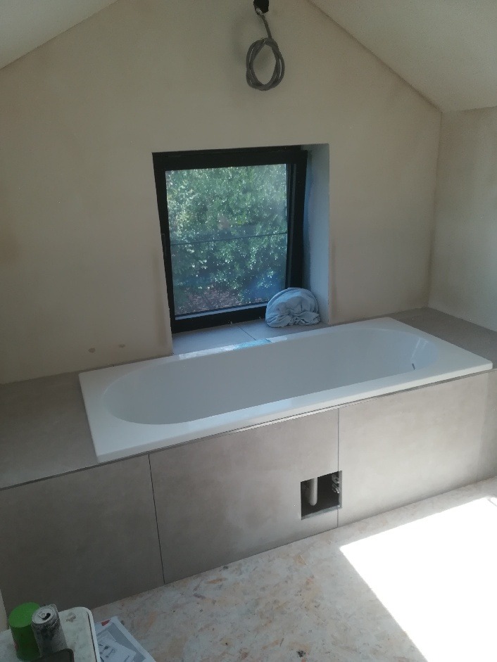 Biała wanna zabudowana szarą płytą, okno z widokiem na zieleń, surowe wykończenie łazienki w trakcie remontu.