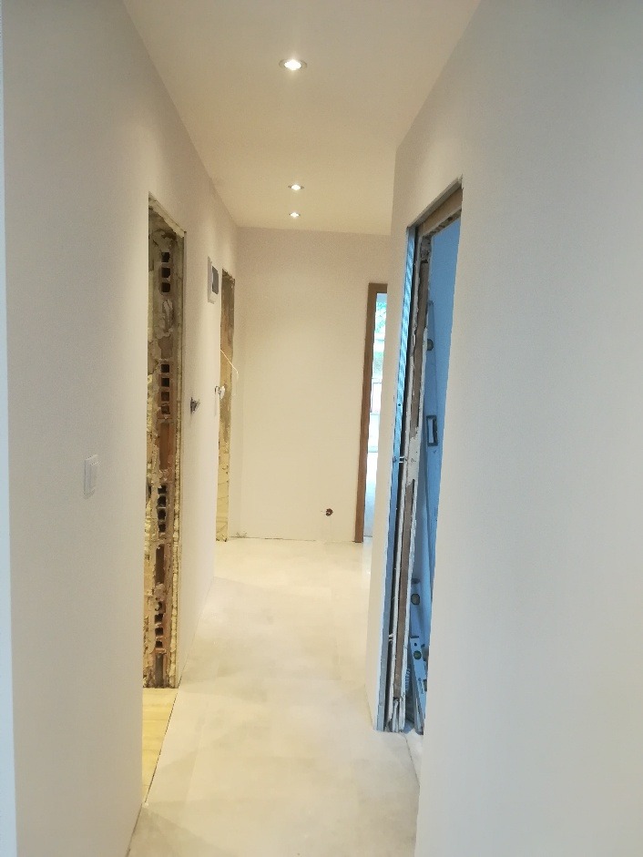 Korytarz w trakcie remontu z widocznymi otworami drzwiowymi w stanie surowym, świeżo pomalowanymi ścianami na biało i zamontowanym oświetleniem punktowym.