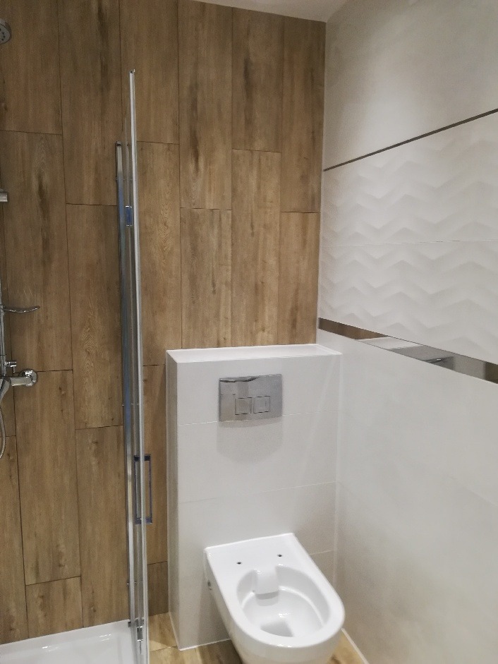 Nowoczesna łazienka z kabiną prysznicową i podwieszaną toaletą, wykończona płytkami imitującymi drewno oraz białymi płytkami z delikatnym wzorem falistym.