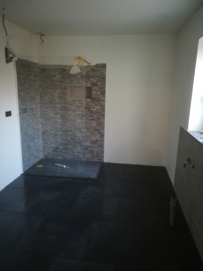 Niedokończona łazienka z czarną podłogą, wykończona szarą mozaiką strefa prysznicowa z brodzikiem i tubą silikonu, białe ściany w trakcie malowania.