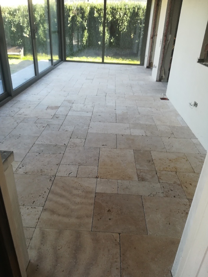 Wykończona podłoga z beżowych, kwadratowych i prostokątnych płytek w jasnym pomieszczeniu z dużymi oknami i widokiem na zieleń na zewnątrz.